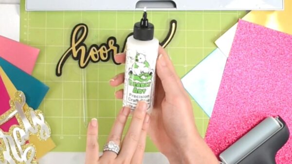 Abbi's hands hold a bottle of Bearly There craft glue over a green Cricut mat, part of a word in vinyl, and the corners of cardstock.