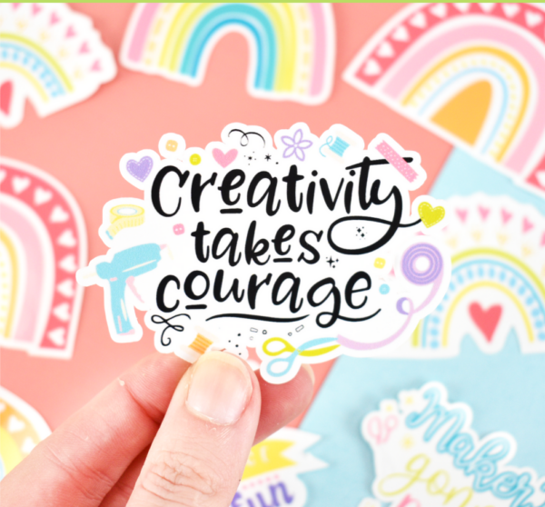 A close-up of Abbi Kirsten's fingers holding a sticker that says, "Creativity Takes Courage." Other printed stickers are on the table below.