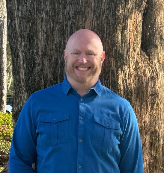 Michael's headshot. He's wearing a blue collard shirt and standing outside, in front of a large tree.