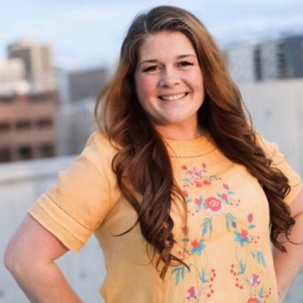 Paula's headshot. She has long brown hair and is wearing a light orange shirt with flowers. She's posing outside.