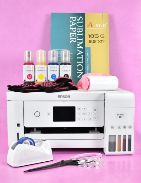 A desktop sublimation printer sits against a plain pink backdrop. Craft rolls and bottles of ink are on top of the printer, and heat safe tape is in a dispenser in the front.