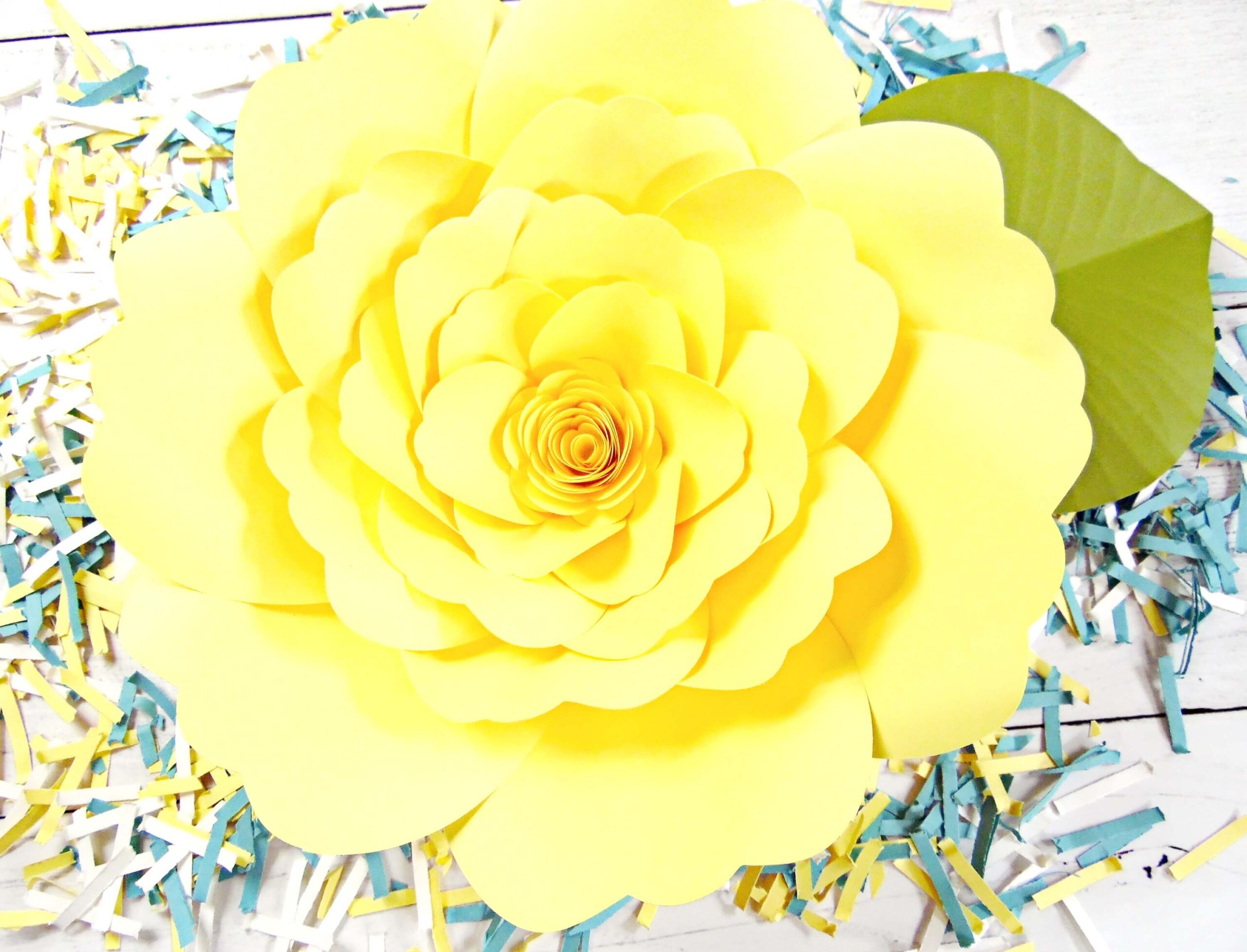 A giant yellow Charlotte paper rose sits on a white background that's covered with confetti strips of yellow, white, and blue paper.