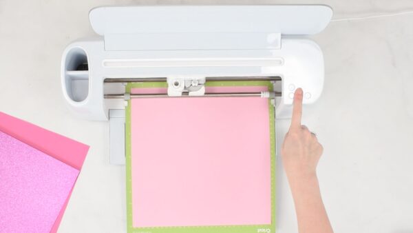 Abbi Kirsten's finger presses the "make it" button on the Cricut machine. There is a mat already loaded with pink paper. 