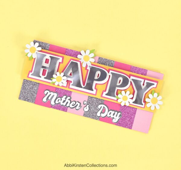 The image shows a pink and purple handmade mother's day card on a yellow background. The card is made with Cricut. 