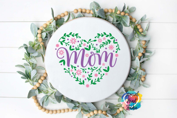 The image shows a floral Mom SVG cut file in pink and purple on a white wood sign. Get this free SVG file here.