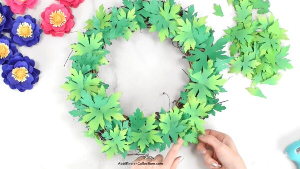 The image shows how to add paper leaves to a grapevine wreath base. 