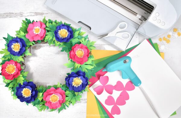 The image shows a paper flower wreath next to a Cricut. Get ready for spring with this fun and easy-to-follow DIY paper flower primrose spring wreath tutorial! Learn how to make it with your Cricut in just a few simple steps.