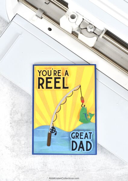 The image shows a DIY Father's Day card on a Cricut machine made from cardstock and iron on vinyl. The card reads, You're A Reel Great Dad. 