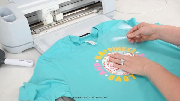 The image shows a blue t-shirt with a Cricut machine and a crafter applying patterned heat transfer vinyl to the shirt. 