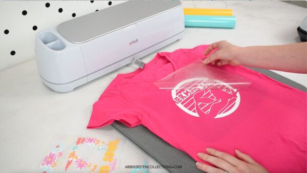 The image shows a pink t-shirt with a crafter peeling off the clear liner carrier sheet to the iron-on vinyl design that has been applied to the shirt. 