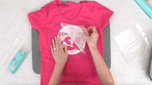The image shows a pink t-shirt with a crafter peeling off the clear liner carrier sheet to the iron-on vinyl design that has been applied to the shirt for the second layer. 