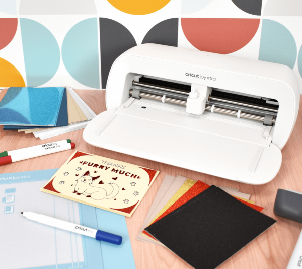 The Cricut Joy Xtra machine on a desk with crafting supplies.