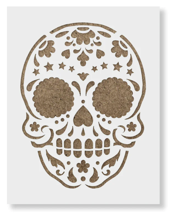 A white stencil of a Dia de los Muertos sugar skull on white paper against a brown cardboard background.