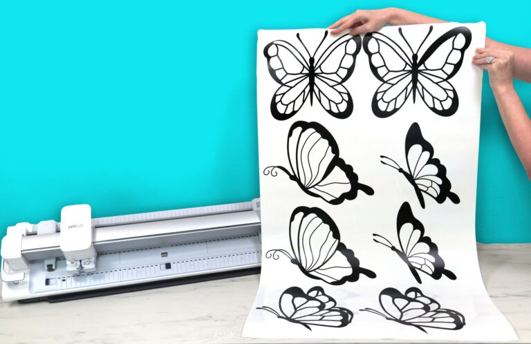 The Cricut Venture die-cutting machine on a white desk with a blue background. Abbi Kirsten's hands hold a large sheet of smart vinyl featuring butterflies that has been cut out on the Venture machine.