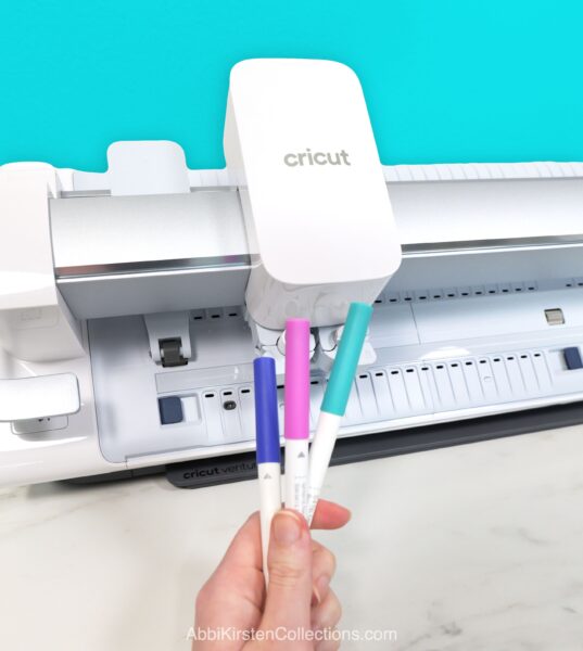 Cricut Venture cutting machine with woman's hands holding Cricut pens in several colors. 