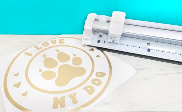 The Cricut Venture cutting machine on a white desk with a blue wall behind it. Next to the Venture is a large vinyl cut out that says, I love my dog. 