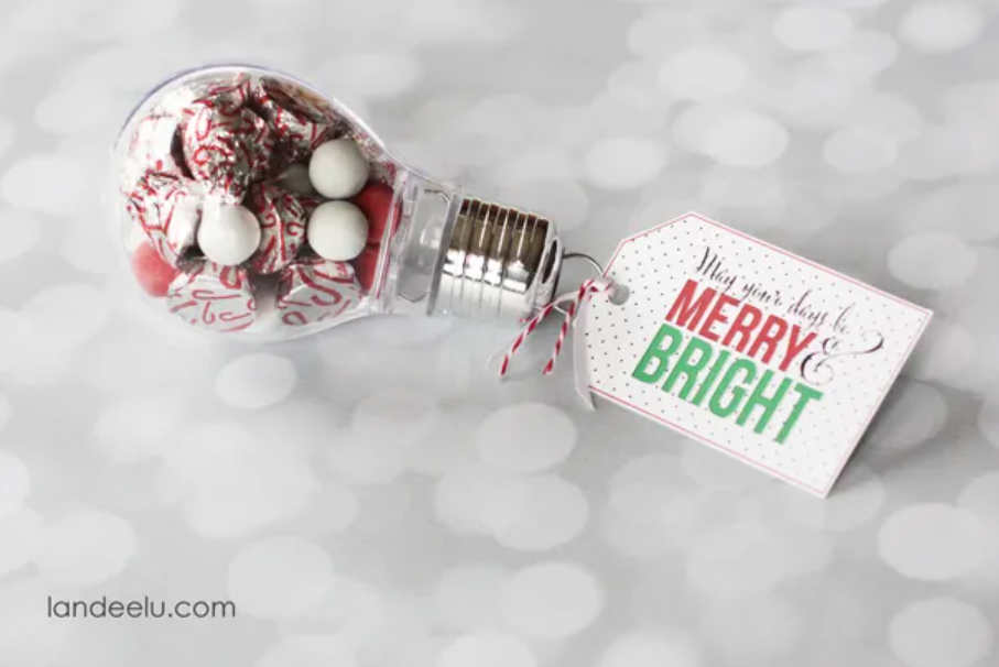 A fake light bulb is filled with Christmas-colored candies. A festive homemade tag on the bulb says, “May your days be merry and bright” in green, red, and gold. The bottom of the picture has the words “Quick and Easy Gift Idea landeelu.com. 