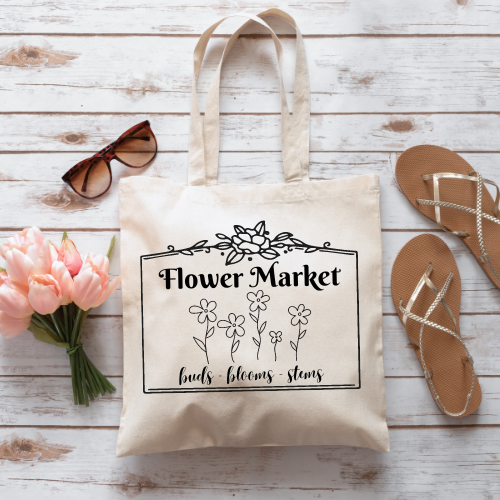 Cream-colored tote bag with text Flower Market, birds, blooms, stems and simple flowers with pink flowers, sunglasses, and sandals.