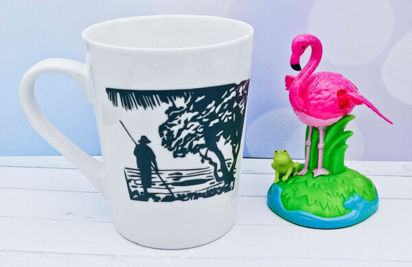 White mug with landscape photo of person on boat and tree with pink flamingo and frog decoration beside mug. 