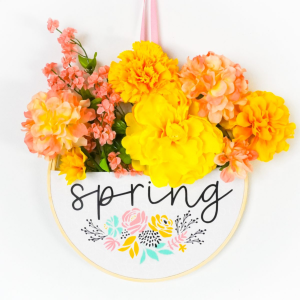 Embroidery hoop wreath with yellow and peach flowers with a SVG cut file that reads Spring added to the front with iron-on vinyl. 