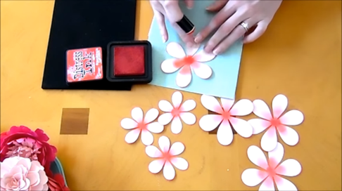 In a screenshot from a ruffle paper dahlia tutorial, Abbi applies pink distress ink to the center of different size paper dahlia petals. She’s working on an orange crafting surface with supplies spread out. 
