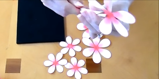 In this video screenshot, Abbi uses a wooden dowl to curl the paper petals of a ruffled dahlia flower. Unrolled petal layers lay on the work surface below her hands. 