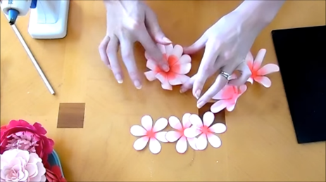 Abbi Kirsten assembles the ruffled dahlia paper flower by gluing petal layers onto each other. Other petal layers, a hot glue gun, and finished paper flowers also lay on the table. 