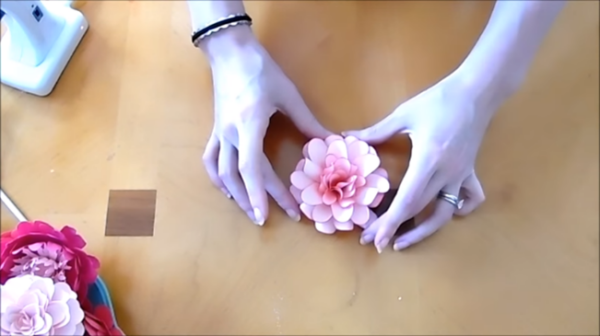 Abbi Kirsten's hands gently cradle a finished ruffled dahlia paper flower on a tan table.