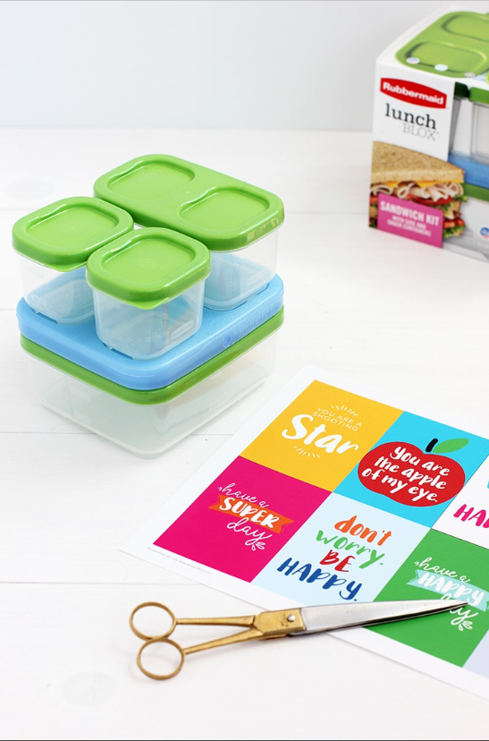 A sheet of lunch box printables lays on a white tabletop next to plastic Rubbermaid lunch box containers and a pair of scissors. The cards have words of encouragement for students, such as “Be Awesome” and “Try Your Best.”